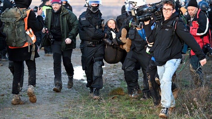 Räumung von Lützerath: Polizisten tragen Greta Thunberg weg, im Vordergrund versucht ein Mann mit TV-Kamera auf der Schulter in eine gute Position zum Filmen zu kommen.