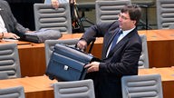 NRW-Finanzminister Marcus Optendrenk wuchtet auf der Regierungsbank einen schweren schwarzen Aktenkoffer über die Sitzreihe. Aufnahme aus der Plenarsitzung am 23.08.2023