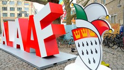 Ein großer rot-weißer Schriftzug "Alaaf" und eine Narrenkappe stehen auf einem Platz in Köln