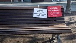 Eine Parkbank mit zwei Schildern - ein rotes mit der Aufschrift "Verweilverbotszone. Bitte gehen Sie weiter", ein weißes mit "Freitag 15 bis 1 Uhr Samstag/Sonntag 10 bis 1 Uhr
