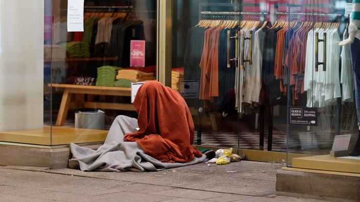 Ein Mann sitzt in der Innenstadt in einem Schlafsack im Eingang zu einem Geschäft. Trotz lebensgefährlicher Kälte meiden manche Obdachlose bereitstehende Notschlafplätze in NRW und harren Wohlfahrtsverbänden zufolge im Freien aus.