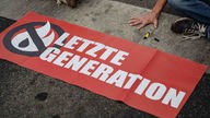 Ein Aktivist der Gruppe Letzte Generation hat seine Hand auf dem Asphalt festgeklebt. Davor liegt ein Banner der Gruppe