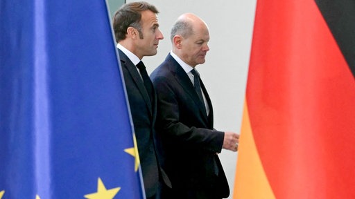 Olaf Scholz und Emmanuel Macron zwischen einer EU- und Deutschland-Flagge.