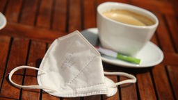 Eine FFP2 Maske liegt neben einer Kaffeetasse auf einem Tisch in einem Café