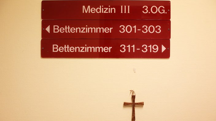 Ein kleines Holzkreuz hängt unter Hinweisschildern auf einer Station in einem kirchlichen Krankenhaus