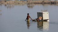 Menschen in Pakistan waten mit Besitztümern durch eine geflutete Landschaft