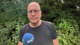 WDR-Reporter Thomas Wenkert steht mit einem Mikrofon in der Hand vor einem Gebüsch