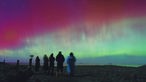 Polarlichter über den Hebriden in Schottland, Menschen stehen an einem Strand  und beobachten das Naturereignis
