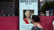 Eine Frau steht vor einer Tafel auf der Queen Elizabeth zu sehen ist 