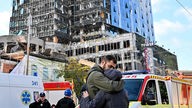 Menschen umarmen sich vor einem teilweise zerstörten mehrstöckigen Bürogebäude