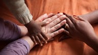 Eine weiße und eine Schwarze Frau und ein Baby legen ihre Hände übereinander. 