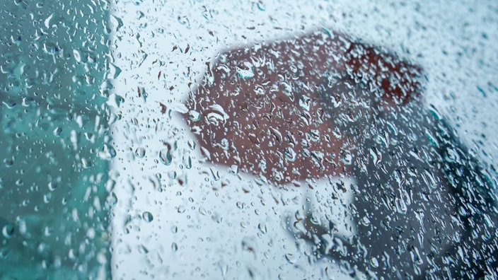 Hinter einer Scheibe mit Regentropfen ist ein Mensch mit Regenschirm zu erkennen.