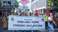 Auf dem Bild sind mehrere Mitarbeitende der Uniklinik Bonn zu sehen, welche auf der Straße mit Fahnen und Bannern demonstrieren. In ihrer Mitte halten sie ein Banner mit der Aufschrift:"Uniklinik Bonn fordert Entlastung".