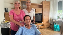 Drei Frauen stehen in einem Zimmer des Pflegeheims. Eine von Ihnen sitzt in einem Rollstuhl und ist eine Bewohnerin der Pflegeeinrichtung.