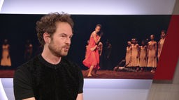 Der Intendant des Tanztheaters Wuppertal, Boris Charmatz, erzählt von der neuen Aufführung "Le Sacre". Der Bildschirm im Hintergrund zeigt eine Aufnahme aus der Aufführung: Afrikanische Tänzer bewegen sich in seidenen Kleidern auf einer dunklen Bühne.