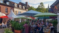 Dutzende Menschen sitzen unter Sonnenschirmen auf einem Platz zwischen Wohnhäusern in der Abendsonne