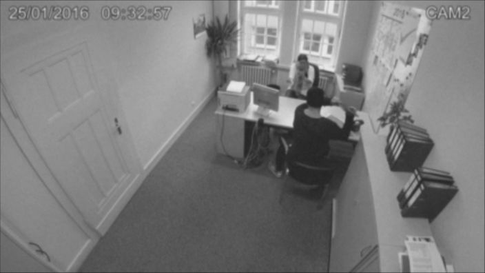 Eine schwarz-weiße Aufnahme einer Sicherheitskamera, welche in einem Büro einer Behörde installiert ist. Die Mitarbeiterin wird von einem Kunden bedroht.