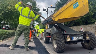 Ein Arbeiter schaufelt Asphalt aus einem Anhänger in die Ausfräsung für die Glasfaserleitungen