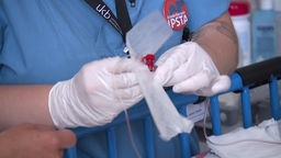 Person in medizinischer Arbeitskleidung und Handschuhen öffnet Katheter. 
