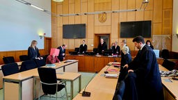 Auf dem Foto ist ein Gerichtssal, in dem der Richter gegenüber eines Mannes sitzt, der sich eine Mappe vor sein Gesicht hält.