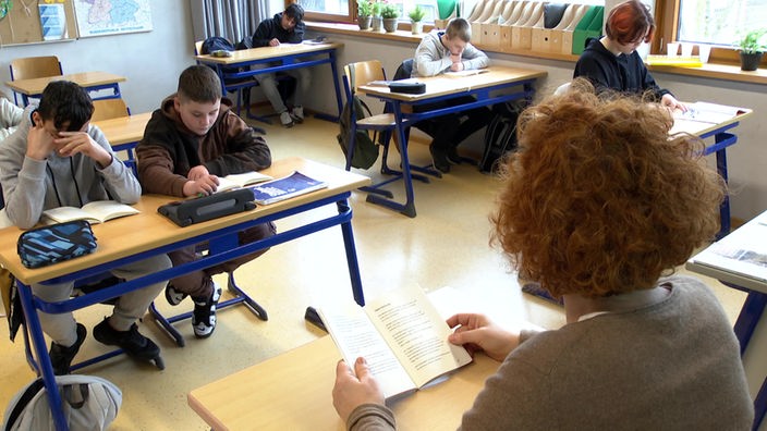 Eine Lehrerin sitzt mit einem aufgeschlagenem Buch vor Schülern in einer Klasse