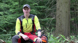 Ein junger Mann in greller Forstwirt-Kleidung sitzt im Wald auf einem Baumstumpf