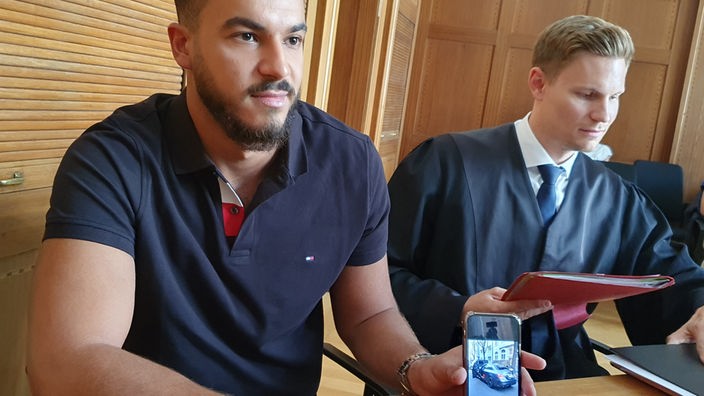 Ein junger Mann sitzt im Gericht und zeigt auf ein Foto von seinem Auto auf seinem Smartphone. Neben ihm sitzt sein Anwalt. 