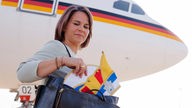 Flugzeug weiß und schwarz-rot-gold lackiert davor Annalena Baerbock mit Handtasche und Narrenkappe
