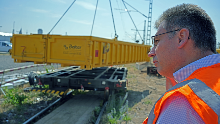 Ein Mann steht in orangener Warnweste vor einem Gleiswagen auf Schienen.