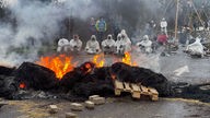 Ein Straßenbarrikade brennt, im Hintergrund sitzen mehrere Menschen mit weißen Overalls und Masken