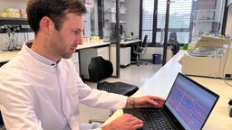 Auf dem Bild ist ein der Forscher Axel Schmidt vom Institut für Humangenetik an der Uniklinik Bonn zu sehen. Vor ihm steht ein Laptop. Auf dem Bildschirm sind bunte Linien zu sehen, die Beethovens Erbgut darstellen.