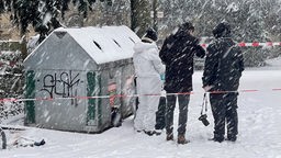 Drei Polizisten stehen in einer winterlichen Landschaft neben Müllcontainern; Einer trägt eine Kamera mit sich