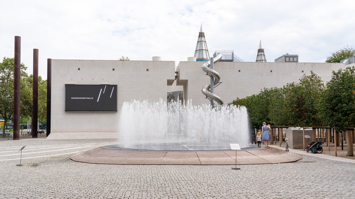 Man sieht die Fassade der Bundeskunsthalle Bonn. Davor ist ein Springbrunnen, auf dessen Rand eine Frau mit Kindern spaziert.