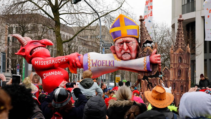 Auf dem Foto ist ein Karnevalswagen, auf dem eine Figur, die Kardinal Woelki darstellt, sich an einen Kirchturm klammert. Dahinter steht ein Teufel mit der Aufschrift "Missbrauchsskandal", der den Karndinal an den Füßen wegziehen will.