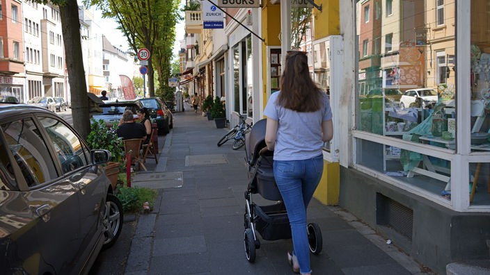 Auf einem Gehweg zwischen parkenden Autos links und Geschäften rechts geht eine Frau mit einem Kinderwagen entlang.