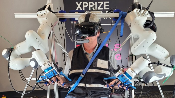 Zu sehen ist ein Mann aus der Jury, der den Roboter testet und durch die Aufagebn manövriert. Er trägt eine VR-Brille und seine Hände und Füße stecken in Bedienelementen.