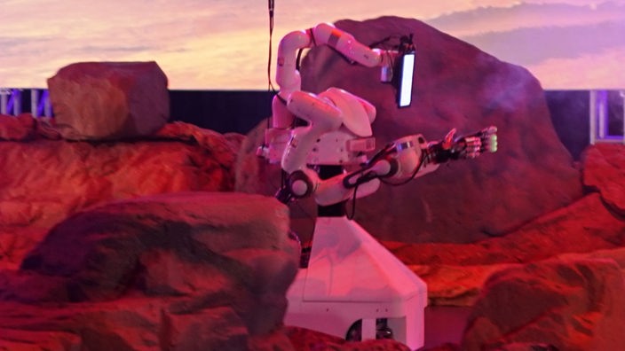 Zu sehen ist der Roboter des Bonner Teams NimbRo in einer rötlich ausgeleuchteten, felsenartigen Wettbewerbsumgebung.