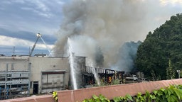 Das Gebäude von außen, Rauch steigt auf und Wasserstrahler versuchen zu löschen