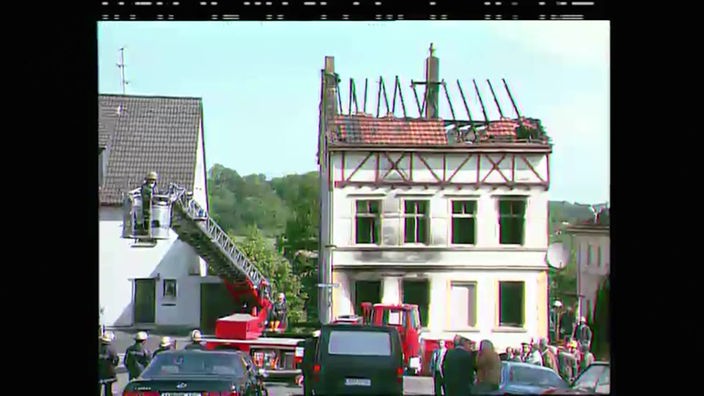 Ein Gebäude mit ausgebranntem Dachstuhl und davor stehen Feuerwehrautos und mehrere Menschen.