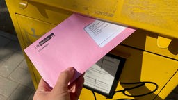 Eine Hand steckt einen rosa Wahlbrief in einen Briefkasten