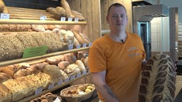 Bäcker Mark Grania hält ein Brot in seinen Händen, im Hintergrund die Backwaren-Auslage.