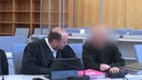 Zwei Personen sitzen in einem Gerichtssaal. Es handelt sich um einen Anwalt und einen Angeklagten, das Gesicht des Angeklagten ist verpixelt.