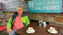 Bernd-Uwe Mach ist Mitgründer des Genossenschaftsprojekts und sitzt mit Kaffee draußen an Tisch 12. Im Hintergrund ist ein türkises Schild mit der weißen Schrift "Café und Bistro Hauderei".
