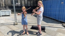 Julia und Lea spielen Geige, hinter ihnen ein Zaun einer Baustelle