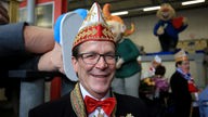 Christoph Kuckelkorn lächelnd und gekleidet als Zugleiter, im Hintergrund die Halle der Festkomitees des Kölner Karnevals 