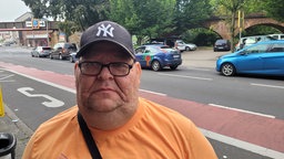 Ein Mann mit kräftigem Gesicht, Kappe und Brille vor einer Straße, auf der mehrere Autos vorbeifahren