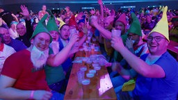 Mehrere Fans sitzen bei der Darts-Meisterschaft am Tisch und trinken aus Bechern