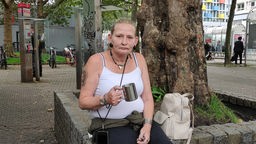 Auf dem Foto ist Sandra Burkhardt, die mit einem silbernen Becher in der Hand auf einer Steinmauer sitzt.