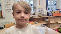 Ein neunjähriger Junge mit blonden Haaren und hellem Pullover in einem Klassenzimmer