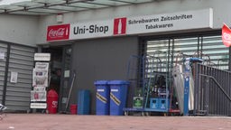 Auf dem Foto ist der Eingang zu einem Kiosk: Eine kleine Tür mit Zeitungsständer daneben, auf einem Schild steht "Uni-Shop".